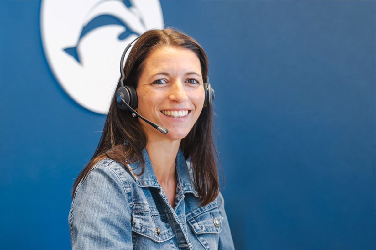 Lächelnde ORCA-Mitarbeiterin mit Headset vor blauem Hintergrund mit ORCA-Logo, bereit für den Support-Anruf